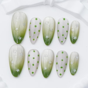 May include: A set of ten almond-shaped press-on nails featuring a gradient design transitioning from white to green. Some nails display a pattern of small green polka dots, while others feature vertical silver glitter lines accented with white star shapes.