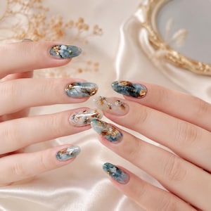 Könnte beinhalten: Oval geformte Fingernägel mit einem Marmordesign in Blau-, Grau- und Weißtönen, akzentuiert durch eingebettete Blattgoldflocken. Die Nagelkunst zeigt ein glattes, glänzendes Finish mit variierenden Mustern aus Farbwirbeln und Goldakzenten.