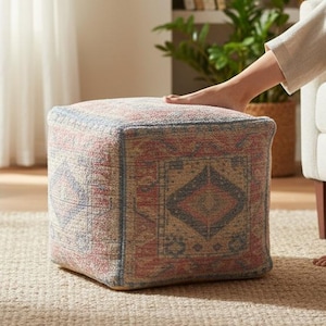May include: A patterned square ottoman in red, blue, and beige tones. The ottoman is placed on a neutral rug in a living room setting. A person is seated in a cream armchair, with their feet resting on the ottoman.