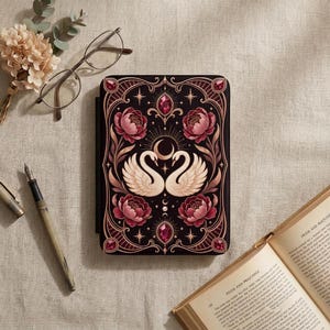 Könnte beinhalten: Eine rechteckige E-Reader-Hülle mit schwarzem Hintergrund und einer symmetrischen Illustration von zwei weißen Schwänen, die sich unter einer Mondsichel gegenüberstehen, umgeben von rosa Pfingstrosen, goldenem Filigran und dunkelroten Edelsteinakzenten.