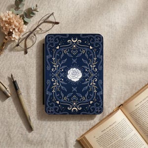 Könnte beinhalten: Ein marineblaues Notizbuch mit einem verzierten Design mit einer weißen Rose, Sternen und Monden. Der Einband ist von goldenen und weißen floralen Mustern umgeben. Ein Füllfederhalter, eine Brille und ein Buch sind ebenfalls zu sehen.