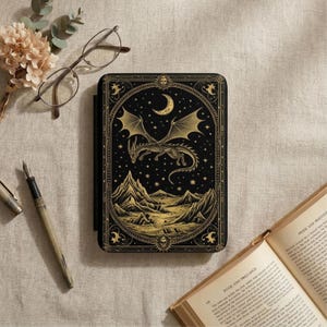 Könnte beinhalten: Schwarzes Notizbuch mit einem goldenen Drachen- und Bergdesign. Das Cover zeigt einen Drachen mit ausgebreiteten Flügeln, einen Mond und Sterne. Ein Stift, eine Brille und ein Buch sind ebenfalls zu sehen.