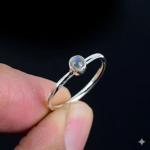 May include: A thin silver ring featuring a small, round, iridescent gemstone set in a simple bezel. The metal band has a polished, reflective finish, and the stone displays subtle blue and grey tones under the light, creating a delicate appearance.