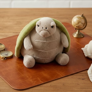 Puede incluir: Un peluche de tortuga con un caparazón verde claro y una expresión triste. La tortuga es de color gris claro y está sobre una alfombrilla de escritorio de cuero marrón. Un pequeño globo terráqueo y una brújula están en el fondo.