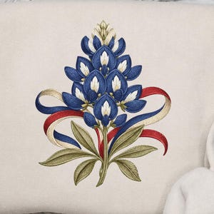 May include: A cream-colored sweatshirt featuring a central embroidered design of a bluebonnet flower in shades of blue and white, accented with green leaves and a flowing red, white, and blue ribbon wrapped around the base of the floral stem.