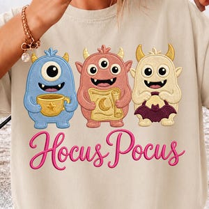 Peut inclure: Un t-shirt beige avec une broderie représentant trois monstres colorés. Le monstre bleu tient une tasse dorée, le monstre rose tient un parchemin et le monstre crème tient une chauve-souris violette. Le texte Hocus Pocus est brodé en rose.