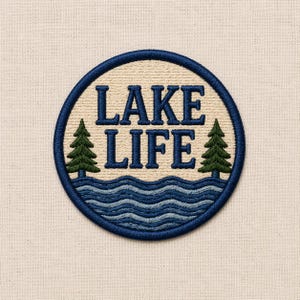Op de afbeelding: Geborduurde ronde patch met de woorden "LAKE LIFE" in marineblauw. Het ontwerp bevat blauwe watergolven en twee groene dennenbomen. De patch heeft een marineblauwe gestikte rand. De achtergrond is een lichtbeige stof.