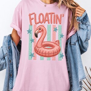 Puede incluir: Camiseta rosa claro con un gráfico de flotador de flamenco. El diseño incluye la palabra "FLOATIN'" en letras naranjas sobre un flotador de flamenco rosa. El fondo tiene rayas verdes y blancas con acentos de estrellas azules. Una camiseta informal con temática de verano.
