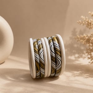 Op de afbeelding: Een stapel van zes ronde armbanden op een rij. Twee effen witte armbanden vormen de buitenste randen, terwijl de vier binnenste armbanden een wervelend gemarmerd patroon hebben in tinten olijfgroen, zwart en wit met een golvend ontwerp.