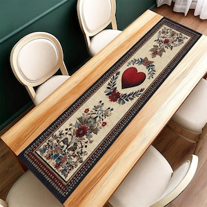 May include: A cream-coloured rectangular table runner with a floral and heart design. The runner features a large red heart in the centre, surrounded by a wreath of leaves and flowers. The edges are bordered with a dark blue pattern.