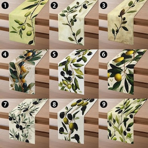 May include: A collection of nine table runners featuring olive branch designs. Each runner displays a different pattern of olives and leaves in shades of green, yellow, and black against various background colors, including cream and beige. The runners are displayed on a wooden surface.