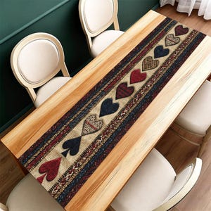 May include: A rectangular table runner with a repeating pattern of hearts and spade suit symbols. The runner features a mix of red, blue, and tan colours with a textured design. The runner is placed on a light wood table.