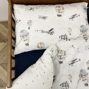 Może przedstawiać: Biały zestaw pościeli z wzorem samolotów w stylu vintage i balonów na ogrzane powietrze unoszących się wśród chmur. Zestaw zawiera poszewkę na poduszkę i poszwę na kołdrę, idealny do pokoju dziecięcego. Pościel znajduje się w drewnianym łóżeczku.