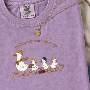 May include: A purple t-shirt with a Comfort Colors label and an embroidered graphic of a mother duck followed by four ducklings. The text "outnumbered by boys" is arched above the ducks. A gold-toned chain necklace rests on the fabric.