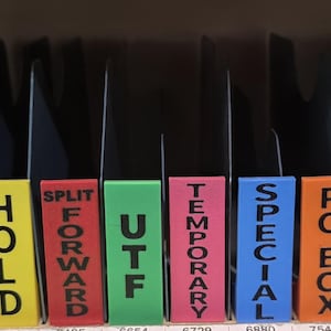 May include: A row of colorful plastic mail sorting dividers with black text labels. The labels read VACATION, DISMOUNT, HOLD, SPLIT FORWARD, UTF, TEMPORARY, SPECIAL, PO BOX, VACANT, and FORWARD. Each divider is a distinct color including orange, pink, yellow, red, green, and blue.