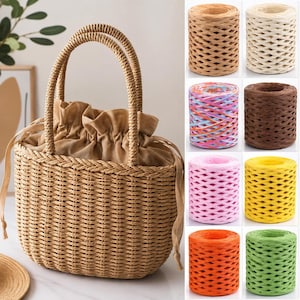 May include: A woven straw handbag with two rounded handles and a tan fabric drawstring closure. Beside the bag are eight cylindrical spools of paper raffia ribbon in various solid colors including beige, white, brown, yellow, pink, orange, and green.