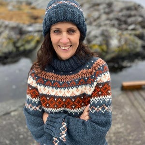 May include: A woman wearing a knitted hat and sweater. The hat and sweater are dark blue with a patterned design in white, orange, and brown. The sweater has a turtleneck and long sleeves. The background is a blurred outdoor scene with rocks and water.