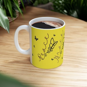 May include: White ceramic coffee mug with a yellow background featuring black script that reads "Peace" and "Joy" with butterfly illustrations. The mug is filled with dark coffee and sits on a wooden surface.