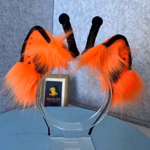 Puede incluir: Una diadema con dos orejas de piel sintética de color naranja brillante con bordes negros y dos antenas negras verticales con puntas redondeadas. Las orejas son esponjosas y texturizadas, colocadas simétricamente en la fina diadema negra.