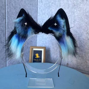 Puede incluir: Una diadema con dos orejas de animal puntiagudas de piel sintética. Las orejas son principalmente negras con acentos de piel en degradado azul y blanco en las secciones internas. Cada oreja está adornada con un pequeño dije de metal plateado.