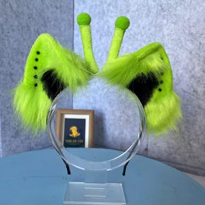 May include: A headband featuring two tall, lime green fuzzy antennae with rounded tips and two large, pointed lime green ears with black fur accents at the base. The ears are decorated with a vertical row of small black dots along the outer edges.