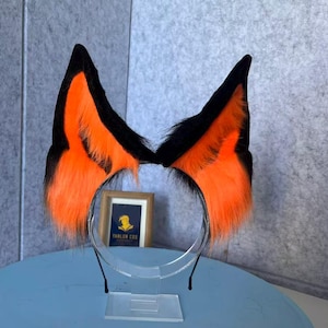 Pode incluir: Uma tiara com duas orelhas de animal pontiagudas feitas de pele sintética preta e laranja brilhante. As orelhas estão presas a uma faixa de metal fina e escura. Uma pequena placa retangular com o texto TANLUN COS é visível entre as duas orelhas.