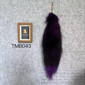 Può includere: Un lungo accessorio a coda in pelliccia sintetica soffice con una sfumatura di colore che passa dal viola scuro in alto al nero al centro, terminando con una punta viola vibrante. Include una clip in metallo per l'aggancio e il testo TMB043.