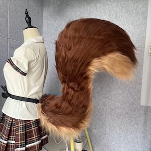Könnte beinhalten: Ein großer, flauschiger braun-beiger Kunstfellschwanz, wahrscheinlich für Cosplay oder Kostüme. Der Schwanz ist an einem schwarzen Gürtel befestigt. Das Fell ist eine Mischung aus dunkelbraun und hellbraun. Der Schwanz ist gebogen und voll.
