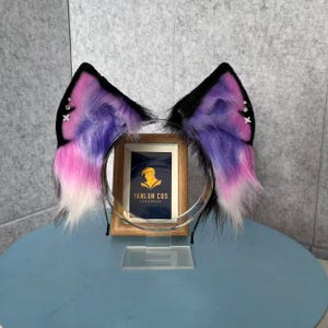 May include: A pair of cosplay cat ears on a black headband. The ears are black with pink, purple, and white faux fur. The ears are decorated with small star-shaped rhinestones. The headband is silver.