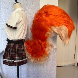 Op de afbeelding: Een etalagepop-torso met een wit overhemd met korte mouwen en een bruine geruite plooirok met een zwarte riem. Aan de taille is een grote, bossige, feloranje en crèmekleurige staart van imitatiebont bevestigd in een gebogen, opstaande vorm.