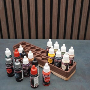 May include: A wooden paint bottle organizer with multiple bottles of acrylic paint in various colors. The bottles are arranged in the organizer and on the surface. The paint bottles have white caps and labels.