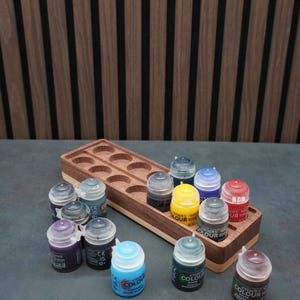 Op de afbeelding: Een houten verfpotjeshouder met een walnootafwerking, met verschillende kleine verfpotjes in verschillende kleuren. De houder heeft cirkelvormige uitsparingen voor elk potje. De achtergrond is een donkere houten wand.