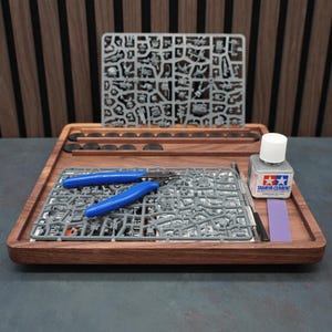 May include: A wooden tray containing hobby supplies. It features plastic model components, blue clippers, Tamiya Cement, and a purple sanding stick. The tray is set against a striped wooden background.