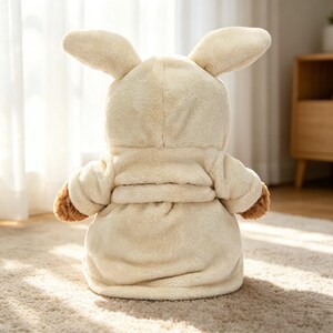 May include: A cream-coloured plush bunny-eared hooded robe. The robe has brown accents on the sleeves and is sitting on a beige carpet. The back of the robe is facing the camera.