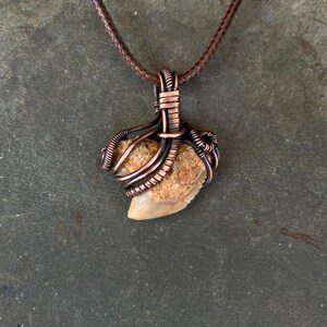 Op de afbeelding: Een hangerketting met een gefossiliseerde haaientand gewikkeld in koperdraad. De tand is lichtbruin van kleur en de draad is donker koperkleurig. De ketting heeft een bruin leren koord.