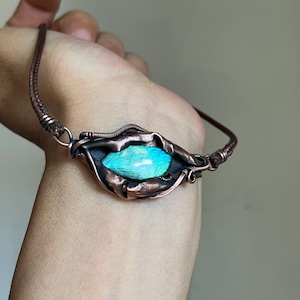 May include: A bracelet featuring a central marquise-shaped labradorite gemstone with iridescent blue and green flashes. The stone is encased in dark, oxidized copper wire wrapping with a textured, organic finish, attached to a braided brown cord.