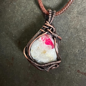 May include: A pendant featuring a raw, translucent white stone with a vibrant pink inclusion, secured in a decorative copper wire wrap. The pendant hangs from a braided copper-colored cord, showcasing intricate metalwork around the stone.