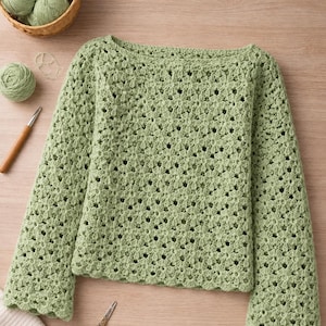 Può includere: Un maglione all'uncinetto verde chiaro a maniche lunghe con un motivo floreale. Il maglione è esposto su una superficie di legno, insieme a un cestino di filato, un uncinetto e delle forbici. Le maniche sono svasate.