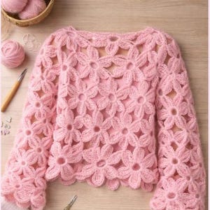 Può includere: Un maglione all'uncinetto rosa chiaro a maniche lunghe con un motivo floreale. Il maglione è realizzato in filato morbido e ha uno scollo tondo. Sono visibili anche un cestino di gomitoli di lana rosa, un uncinetto e delle forbici.