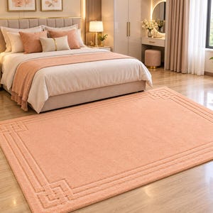 May include: A rectangular peach-colored area rug featuring a textured surface and a decorative border with a geometric Greek key pattern. The rug is placed on a light-colored wooden floor in front of a bed with matching peach-colored pillows.