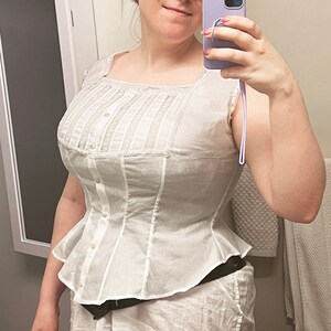 May include: A white cotton corset with a lace trim and button closures. The corset has a fitted bodice and a flared skirt.