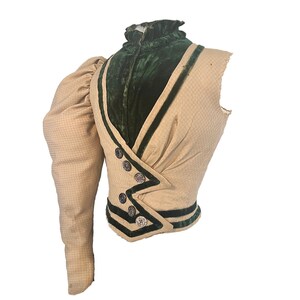 May include: A beige and green vintage waistcoat with a single puffed sleeve. The waistcoat has a high collar and is trimmed with green velvet and black piping. The waistcoat has a button closure and is in good condition.