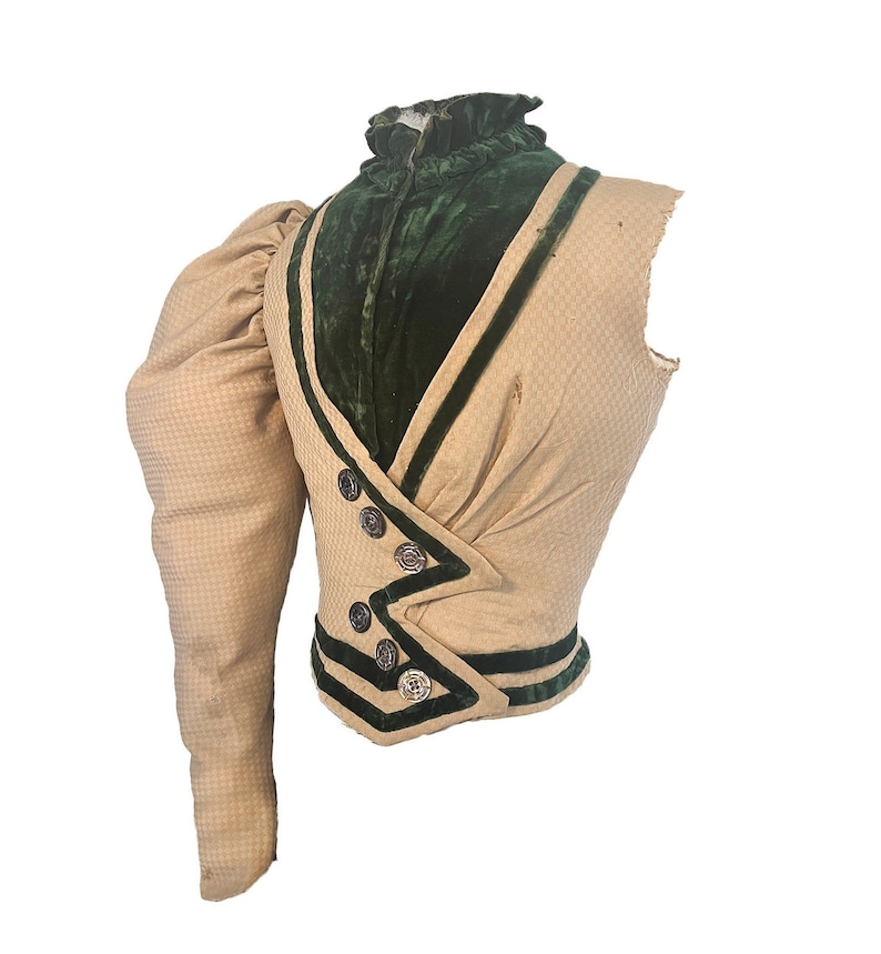 May include: A beige and green vintage waistcoat with a single puffed sleeve. The waistcoat has a high collar and is trimmed with green velvet and black piping. The waistcoat has a button closure and is in good condition.