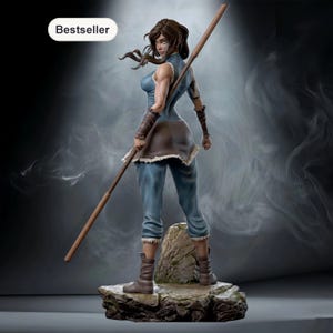 May include: A figurine of a female character in a blue outfit, holding a staff. The figure is standing on a rocky base, with the word "Bestseller" in a white oval in the upper left corner. The background is a smoky gray.