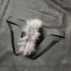 Puede incluir: Un tanga negro con apariencia de cuero sintético texturizado y tiras laterales elásticas finas. Una tira vertical de plumas suaves y esponjosas de color gris claro está fijada en la parte central delantera, creando un contraste de textura.
