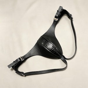 Puede incluir: Un accesorio tipo arnés negro con un acabado liso similar al cuero y un panel central con un patrón texturizado de relieve de cocodrilo. La pieza incluye correas ajustables con hebillas y trabillas de metal plateado para un ajuste seguro.