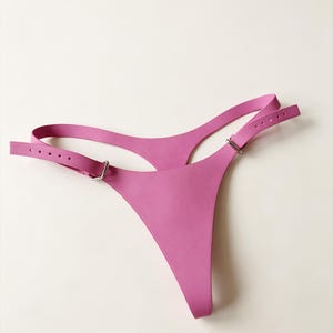 Puede incluir: Una tanga de color rosa brillante con correas laterales ajustables que incluyen hebillas metálicas plateadas y múltiples orificios. La prenda está confeccionada en un material liso de color sólido con un diseño sencillo y corte alto.