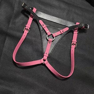 May include: A body harness featuring a central black triangular panel with pink straps attached by silver-toned metal rings and buckles. The design includes adjustable pink bands that connect to a black waistband, all secured with metal rivets and hardware.