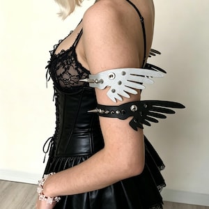 May include: A black leather corset and skirt ensemble. The outfit includes a silver studded choker and arm cuffs with wing-shaped designs. One arm cuff is black, the other is white. The outfit is a gothic or alternative fashion style.
