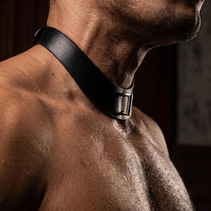 Puede incluir: Un collar de cuero negro con hebilla plateada alrededor del cuello. El collar es grueso y parece estar hecho de cuero de alta calidad. La imagen es un primer plano del collar y la parte superior del cuerpo.